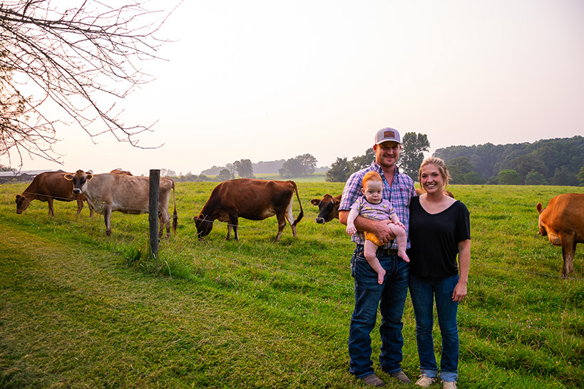 a family of a day, mom, and infant son stands in front of their cows.